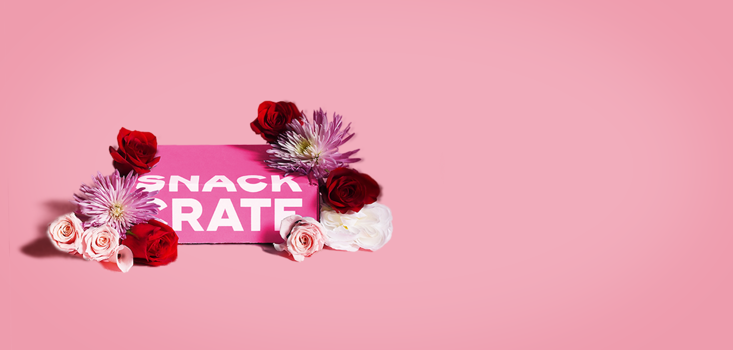 A pink Valentine's Day SnackCrate surrounded by flowers on a pink background